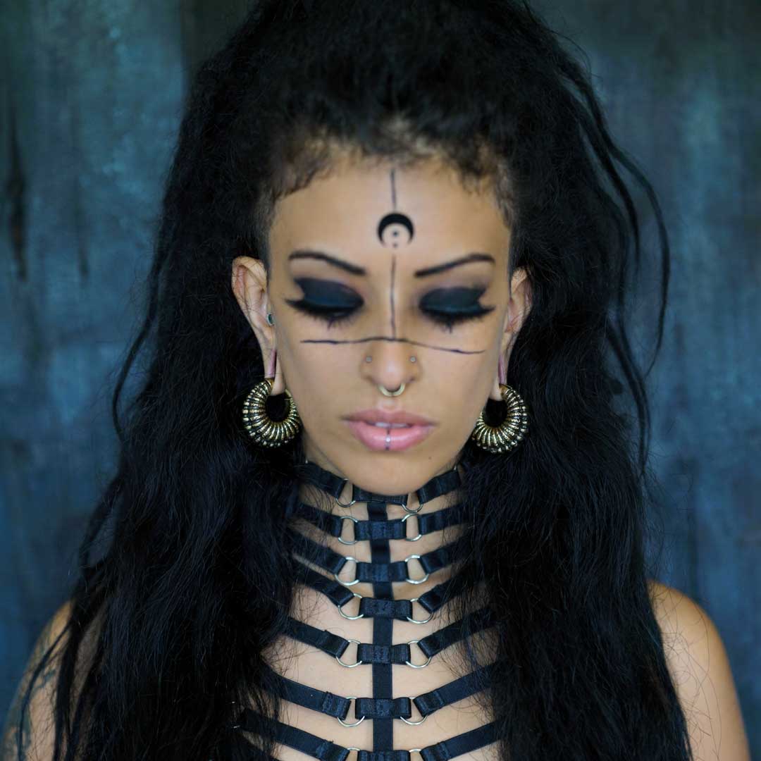 A person with long, dark hair is wearing a harness-style black outfit and the TRILOBITE Large Biomechanical Clicker Ear Weights in Gold Brass by Kaya Movement. Their face features unique makeup that includes a black crescent moon on their forehead, bold eyeliner, and facial piercings. The background is a textured dark blue.
